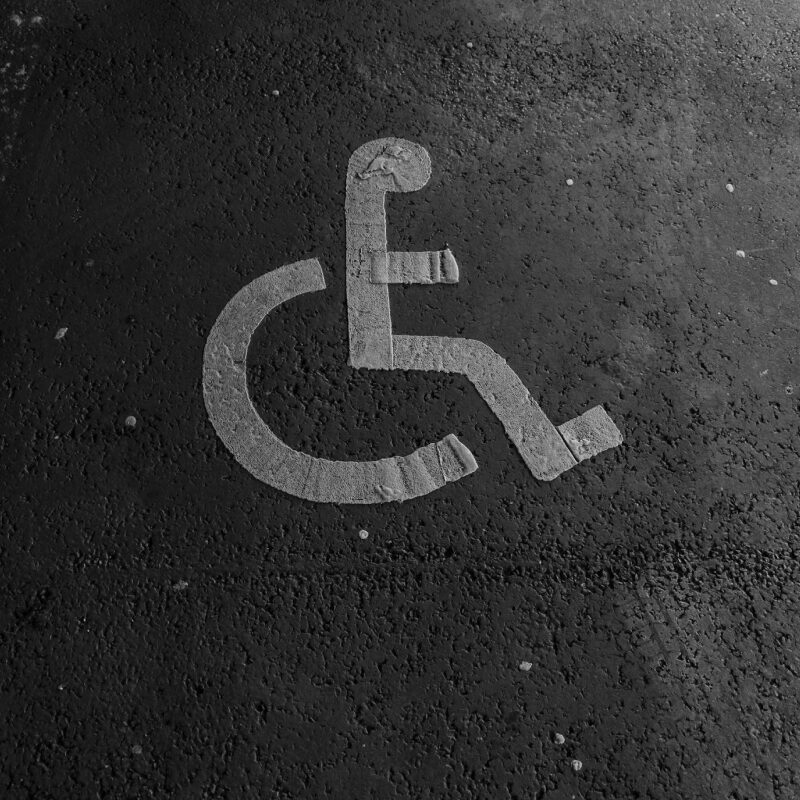 A black and white image of a designated parking spot for people with disabilities. The international symbol of access, a white silhouette of a person in a wheelchair, is painted on the asphalt.