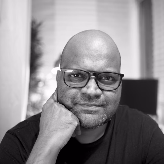 Roshan Ravi - a bald, brown man wearing a black-rimmed glasses and a black t-shirt, smiling at the viewer.