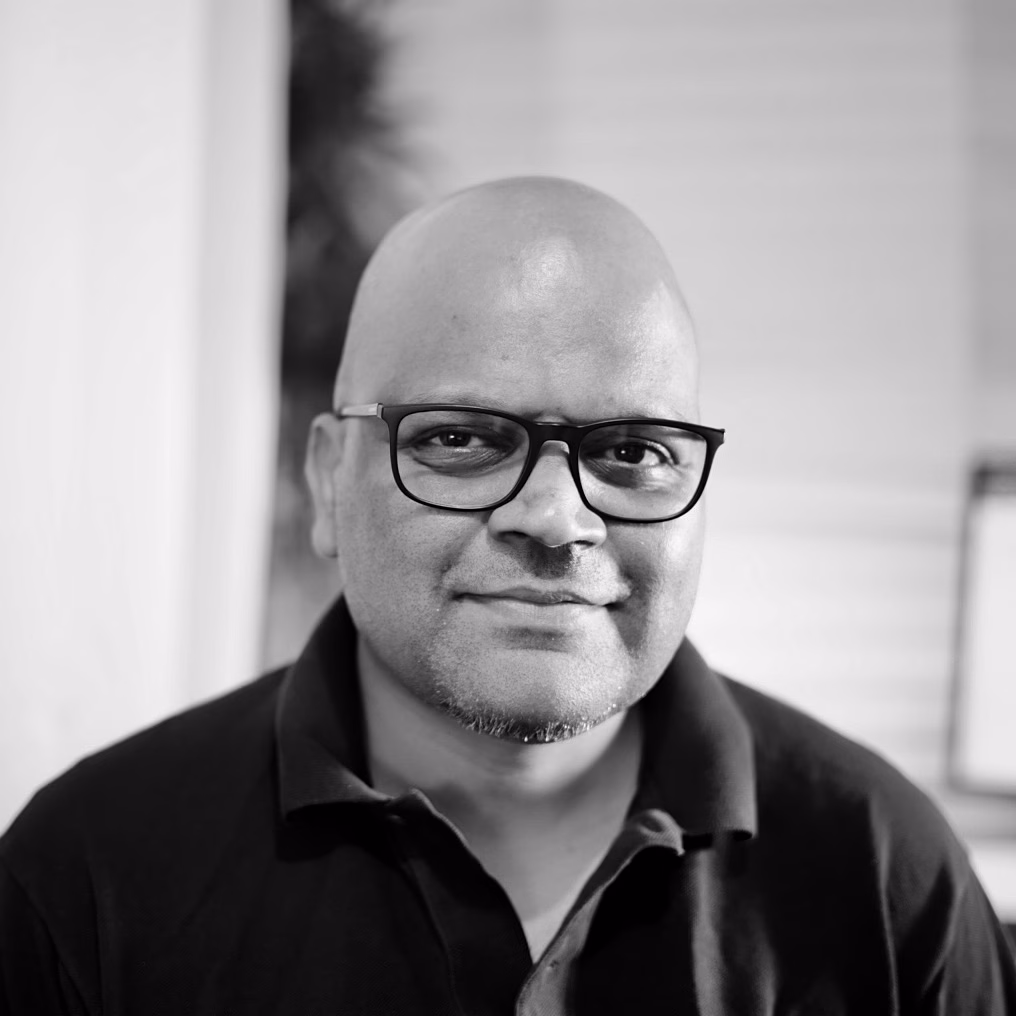 Roshan Ravi - a bald, brown man wearing a black-rimmed glasses and a black t-shirt, smiling at the viewer.